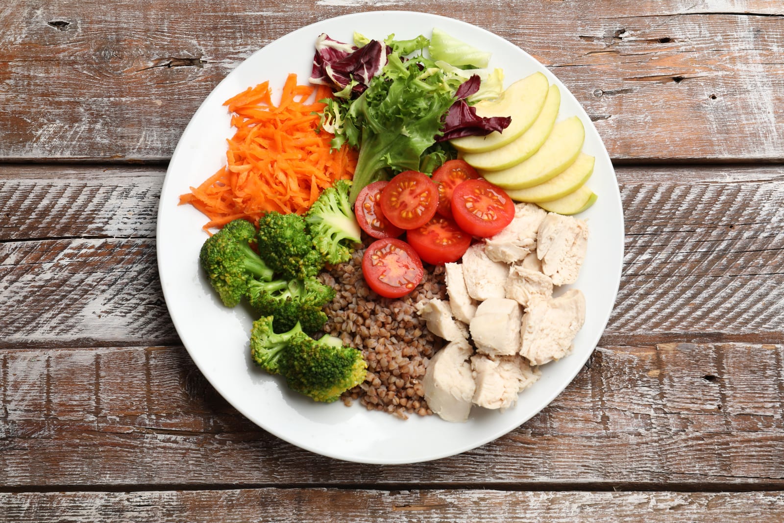 Plato equilibrado con pollo, brócoli, verduras y fruta sobre una mesa de madera