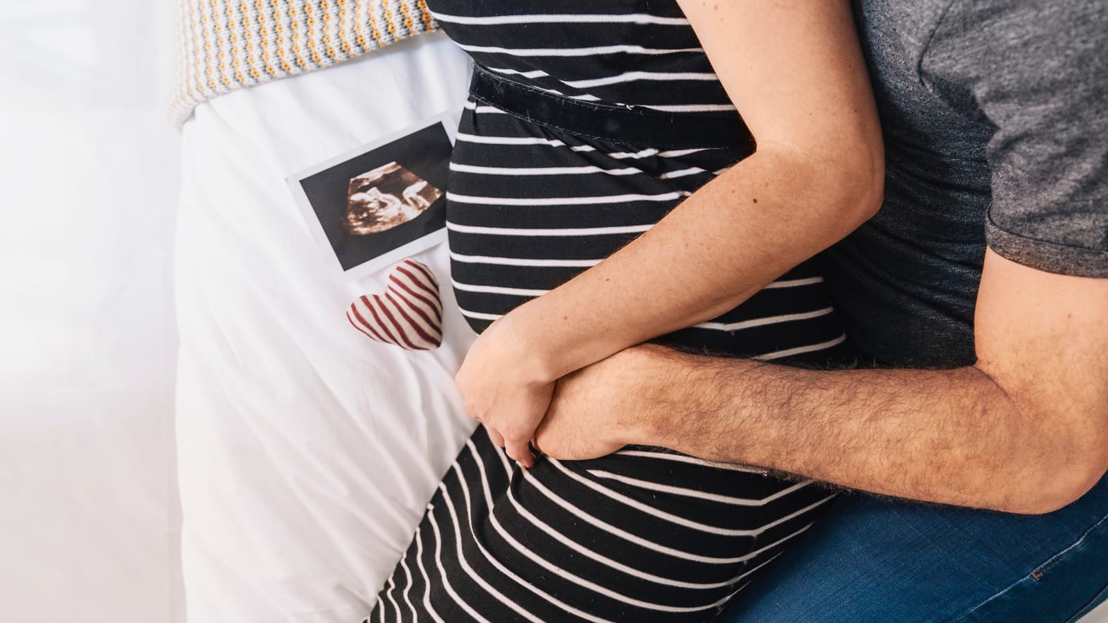 Pareja abrazada junto a un ultrasonido y un corazón de tela sobre la cama